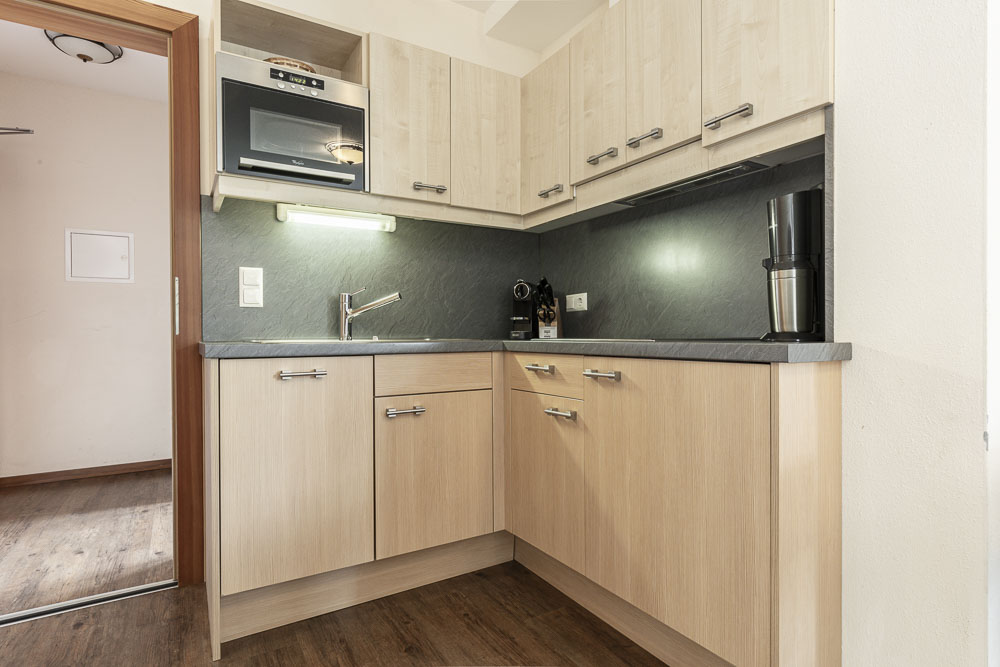 Kompakte Küche im Alpenapart in Ladis, konzipiert für ein Apartment für 4-6 Personen, mit hellen Holzschränken, einer schwarzen Arbeitsplatte und einer kleinen Spüle. Wandmontierte Mikrowelle, Kaffeemaschine und Mühle ergänzen die dunkle Rückwand und den Holzboden für ein gemütliches Ambiente.