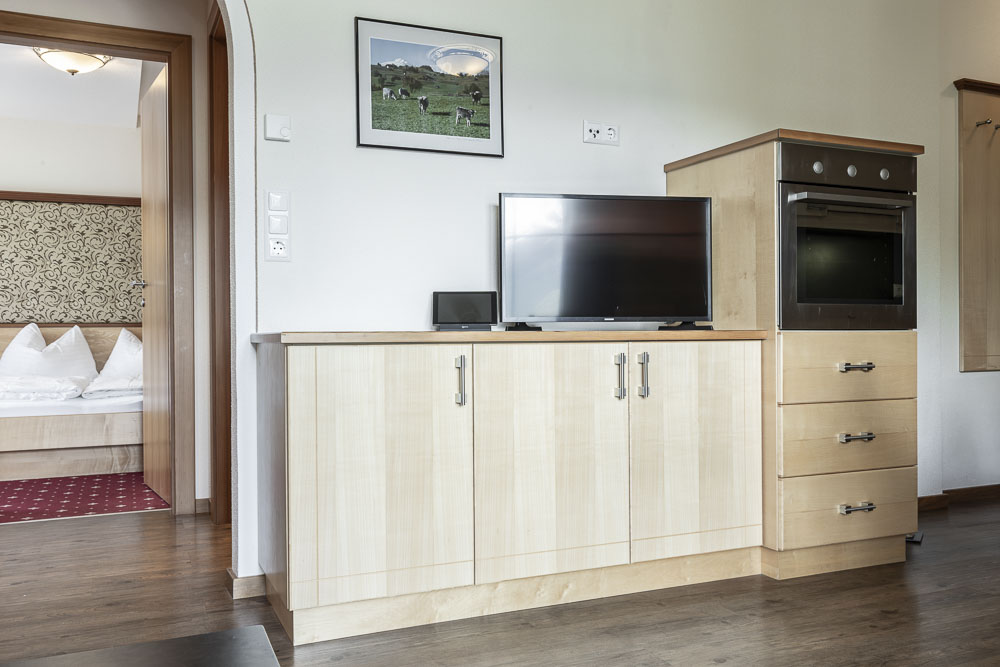 Ein modernes Zimmer im Alpenapart in Ladis verfügt über einen hellen Holzschrank mit Flachbildfernseher und schwarzem Ofen, ergänzt durch ein Kuhfoto an der Wand. Die offene Tür führt zu einem Schlafzimmer mit weißer Bettwäsche und gemustertem Dekor – perfekt für ein Apartment für 4-6 Personen.