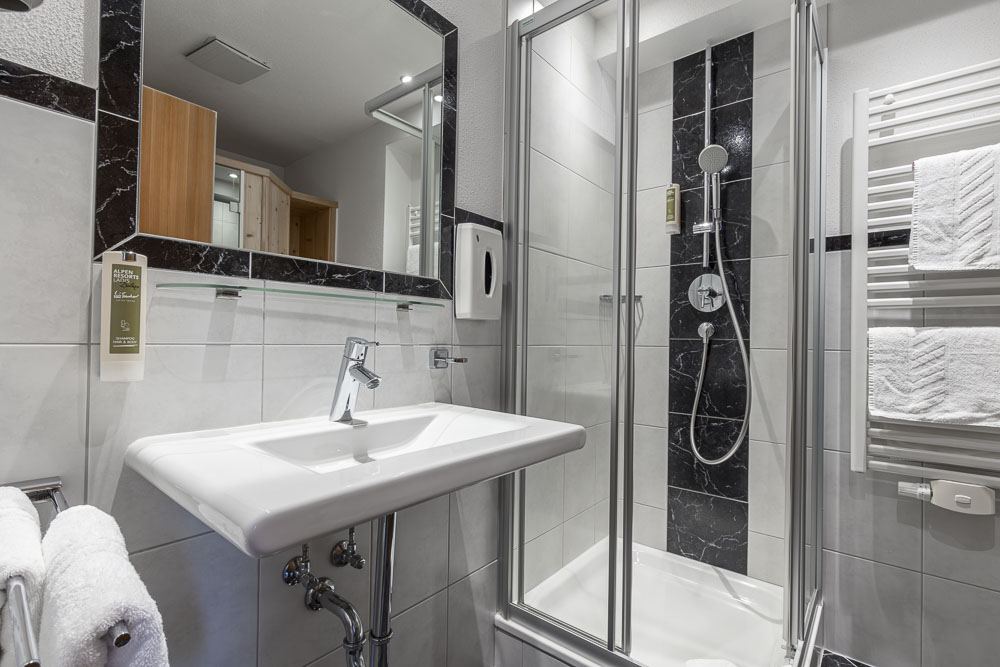 Dieses moderne Badezimmer im Apartment für 4 Personen im Alpenapart in Ladis besticht durch sein elegantes Design. Es verfügt über ein weißes Waschbecken, eine verglaste Dusche mit Regenkopf und Chromarmaturen. Schwarze und weiße Fliesen sorgen für Kontrast, während Handtücher ordentlich am beheizten Ständer neben dem Spiegel hängen.