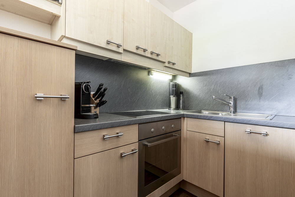 Moderne Küche in einem Alpenapart in Ladis mit Holzschränken, schwarzen Arbeitsplatten und einer Edelstahlspüle. Sie umfasst einen Elektroherd mit Backofen, Messerset und Kaffeemaschine. Helle Beleuchtung unterstreicht das schlichte Design dieses Apartments für 4 Personen.