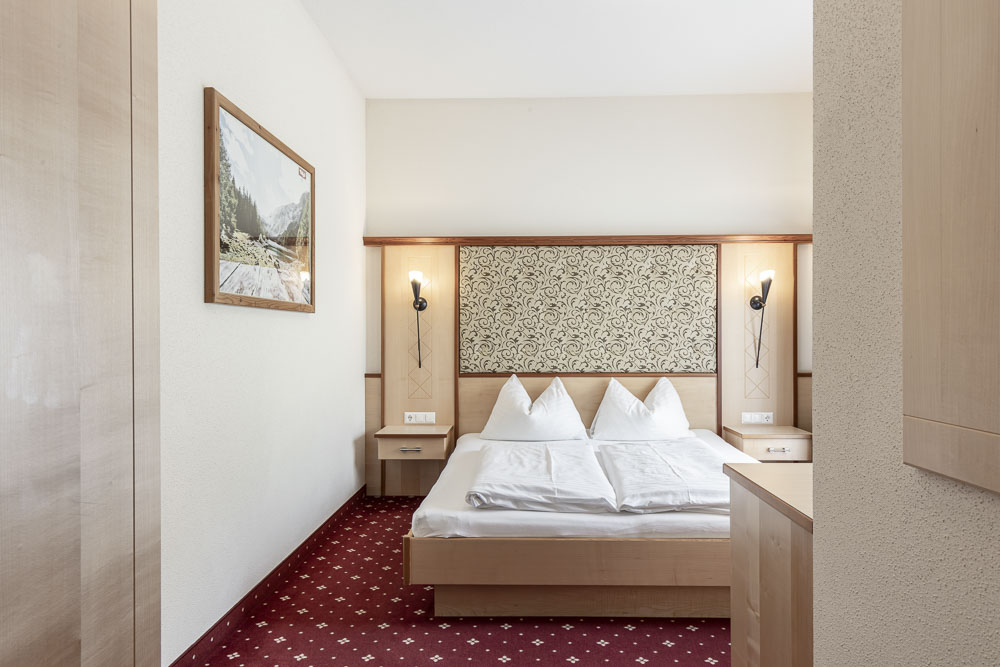 Erleben Sie den Charme eines minimalistischen Hotelzimmers im Alpenapart in Ladis, das zum Entspannen konzipiert ist. Mit einem Doppelbett mit geblümtem Kopfteil, Wandlampen, weißen Wänden und einem gemusterten roten Teppich ist dieser gemütliche Raum perfekt für Ruhe.