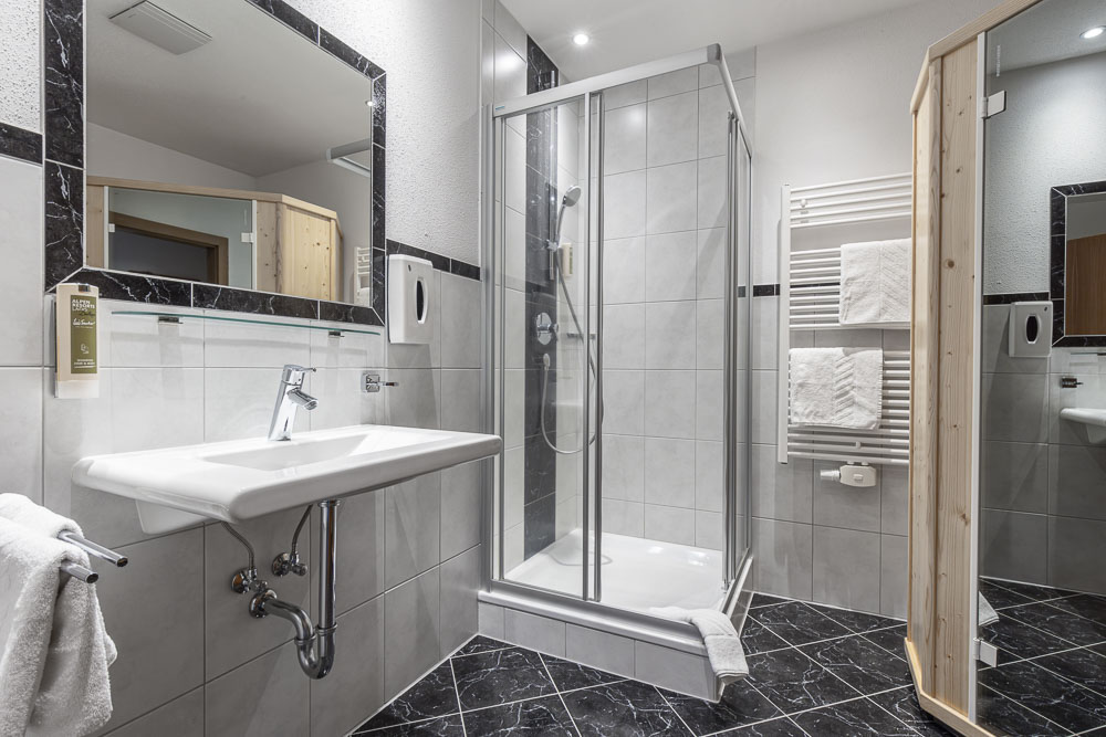 Ein elegantes Badezimmer in einer Wohnung verfügt über eine moderne Dusche mit Glaskabine, ein weißes Waschbecken und einen großen Spiegel. Hellgraue Fliesen schmücken die Wände und bilden einen Kontrast zu den dunklen Bodenfliesen. Ein beheizter Handtuchhalter hält flauschige weiße Handtücher, während auf der rechten Seite eine Holztür teilweise geöffnet ist.