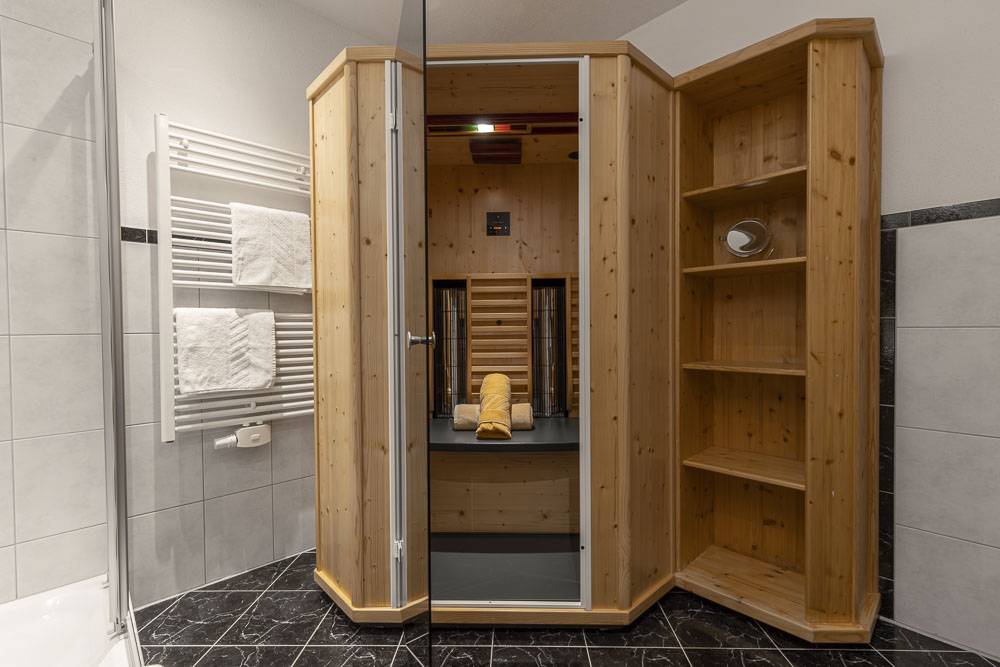 Ein gemütlicher, holzgetäfelter Saunaraum in der Wohnung verfügt über eine Glastür, eine beheizte Bank und ein Bedienfeld. Darin warten ordentlich gefaltete Handtücher auf Sie. Angrenzende offene Holzregale und ein an der Wand montierter Handtuchhalter mit hängenden Handtüchern bieten Komfort über dem dunkel gemusterten Fliesenboden.