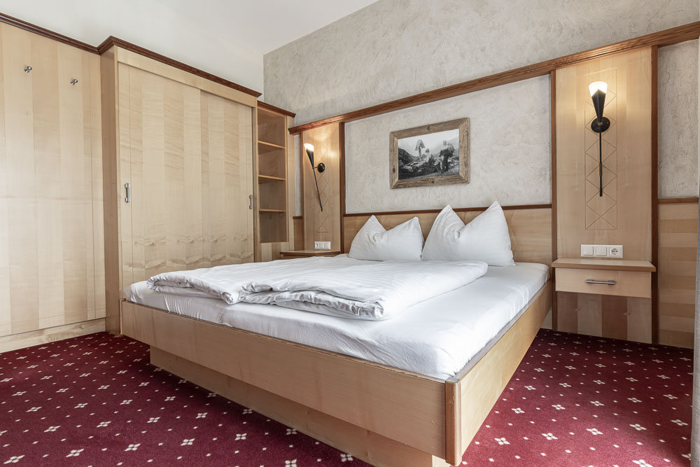 Ein gemütliches Hotelzimmer mit einem Doppelbett aus Holz mit weißer Bettwäsche und Kissen erinnert an die Wärme einer heimeligen Wohnung. Der Raum verfügt über beige Wände, Holzschränke, Nachttische mit Wandlampen und ein gerahmtes Bild über dem Bett. Ein roter Teppich verleiht dem Boden einen Hauch von Luxus.