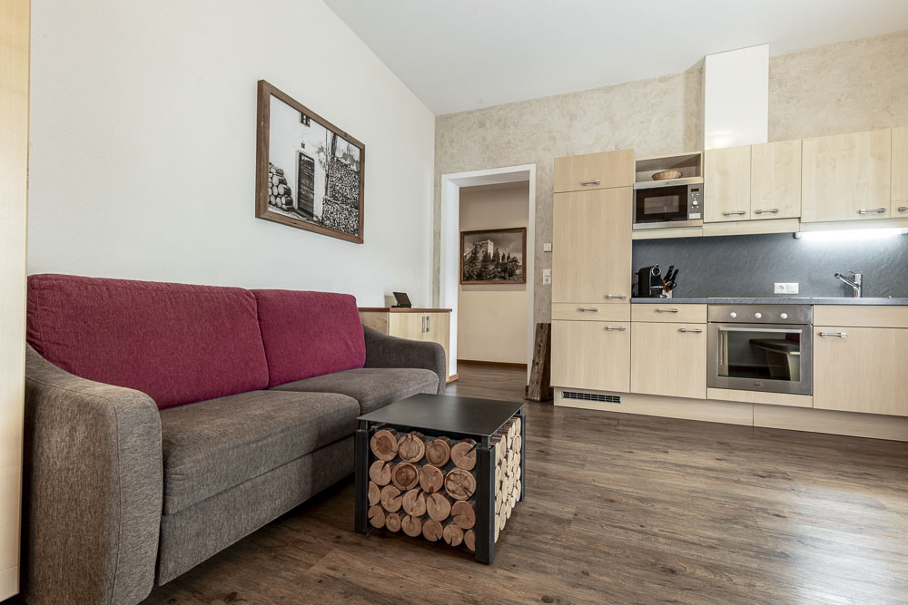 Dieses moderne Apartment verfügt über ein stilvolles Wohnzimmer mit einer grau-roten Couch, einem hölzernen Couchtisch im Blockdesign und einem angrenzenden Küchenbereich mit hellen Holzschränken, einem Ofen und einer Mikrowelle. Ein Schwarzweißfoto schmückt die Wand über dem dunklen Holzboden.