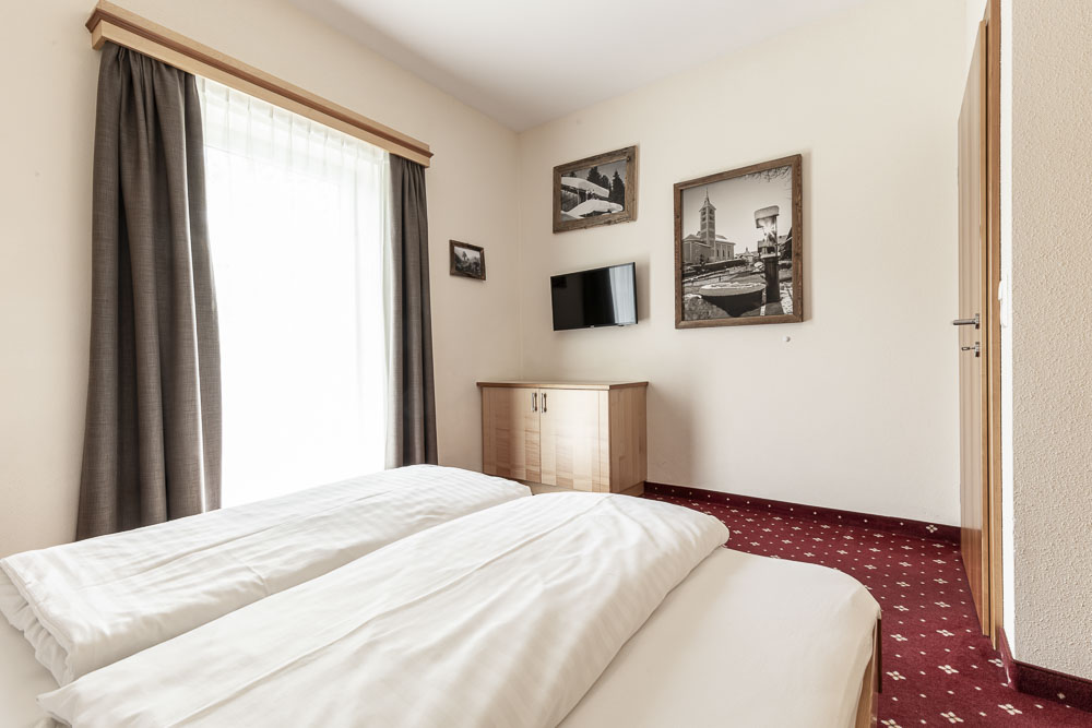 Das Zimmer im Hotelstil des Apartments verfügt über zwei ordentlich gemachte Einzelbetten auf einem roten Teppich. Ein Holzschrank steht unter einem an der Wand montierten Flachbildfernseher, während ein großes Fenster mit durchsichtigen und dunklen Vorhängen den Raum in natürliches Licht taucht. Gerahmte monochrome Bilder verleihen den Wänden eine elegante Note.