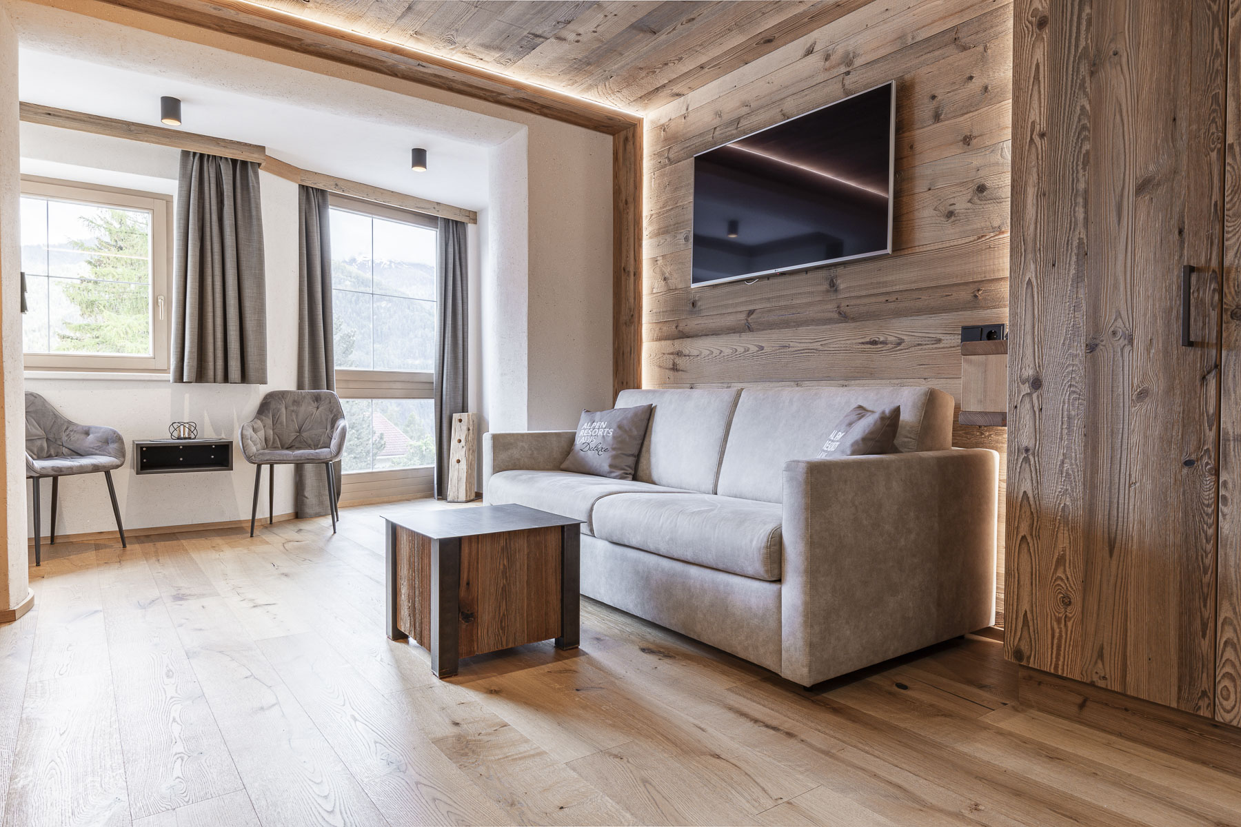 Gemütliches Wohnzimmer mit einem beigen Sofa gegenüber einem an einer holzgetäfelten Wand montierten Fernseher. Helles Tageslicht durch ein Fenster auf der linken Seite, mit zwei grauen Stühlen daneben. Holzboden und ein kleiner Couchtisch verstärken die warme, rustikale Atmosphäre.