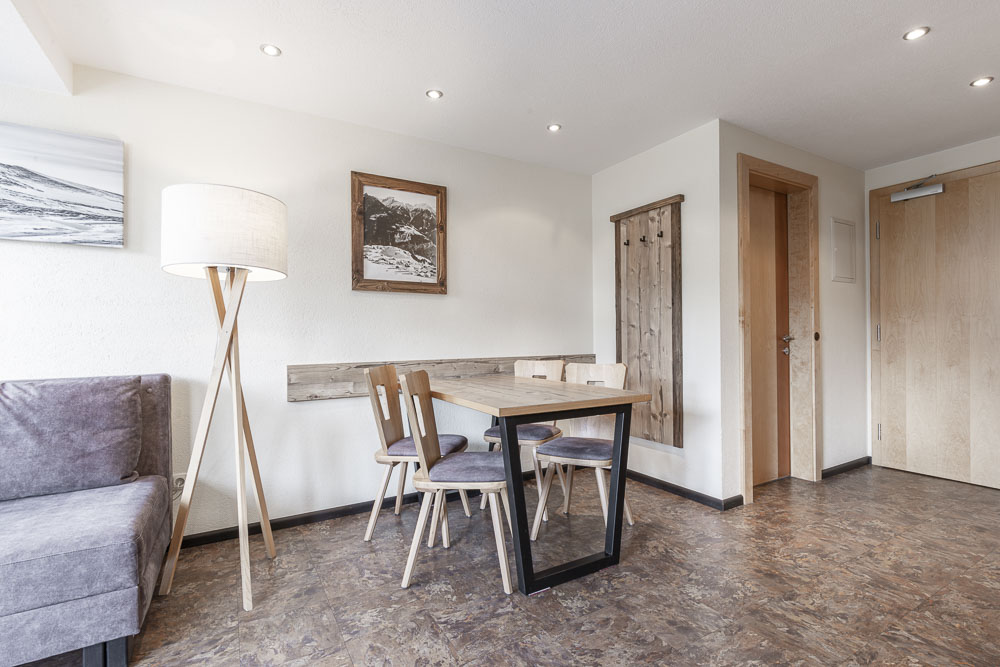 Dieses minimalistische Apartment für 2-4 Personen in Ladis verfügt über einen modernen Holztisch mit vier Stühlen. Eine große Stehlampe steht neben dem grauen Sofa, eingerahmt von weißen Wänden, die mit einem Landschaftsfoto geschmückt sind. Der dunkle Holzboden ergänzt die hölzerne Garderobe neben der Eingangstür.