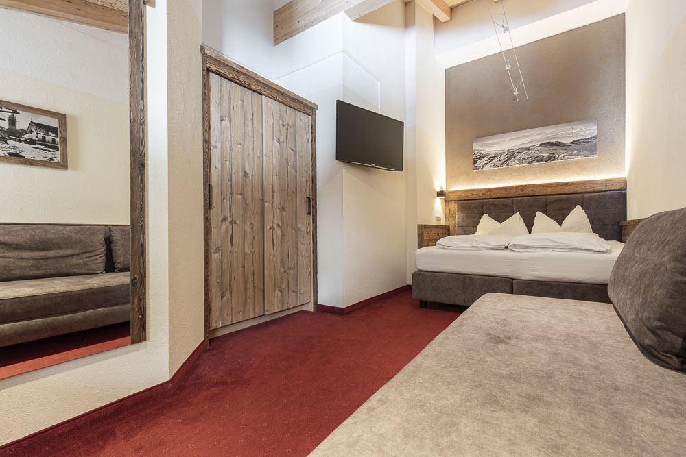 Ein gemütliches Hotelzimmer spiegelt die Ästhetik eines großen Apartments in Serfaus-Fiss-Ladis wider und verfügt über ein Doppelbett, einen roten Teppich und Holzakzente. Ein an der Wand montierter Fernseher und ein schwarz-weißes Landschaftsfoto verleihen diesem malerischen Rückzugsort einen besonderen Reiz.