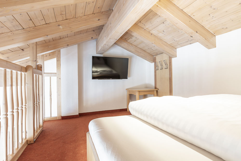 Dieses Schlafzimmer im Loft ist Teil eines großen Apartments für 6-8 Personen in Serfaus-Fiss-Ladis und verfügt über schräge Holzbalken, einen Flachbildfernseher an einer weißen Wand und ein Bett mit weißer Bettwäsche. Es umfasst außerdem einen roten Teppich und ein Holzgeländer sowie einen kleinen Tisch mit Haken unter dem Fernseher.