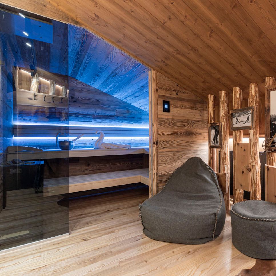 Gemütliches Interieur mit Holzwänden und -decke und einem verglasten Raum im Hintergrund. Zwei dunkle Sitzsäcke und ein runder Ottoman stehen auf einem polierten Holzboden. Sanftes Umgebungslicht verstärkt die warme Atmosphäre.