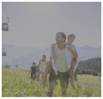 Eine fröhliche Familie wandert durch eine sonnenbeschienene Wiese mit Bergen im Hintergrund, die an malerische Alpenresorts erinnern. Ein Mann trägt ein Kind auf dem Rücken, gefolgt von einem weiteren Erwachsenen und zwei Kindern. In der Ferne links sind Seilbahnen zu sehen.