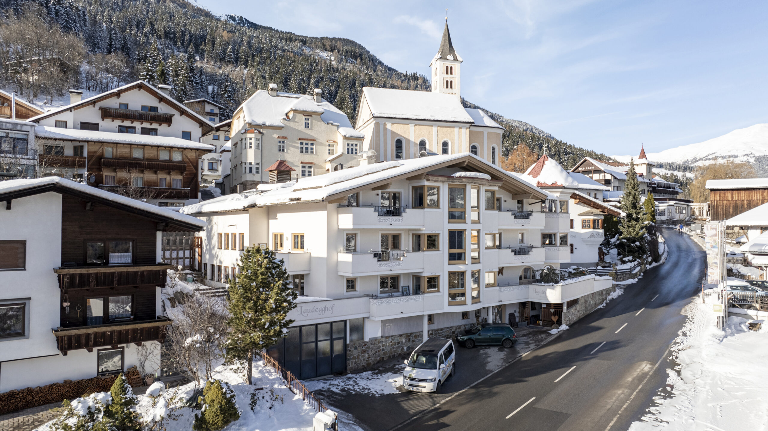 Alpendorf mit verschneiten Gebäuden, Chalet Ladis, einer Kirche und einer kurvenreichen Bergstraße.