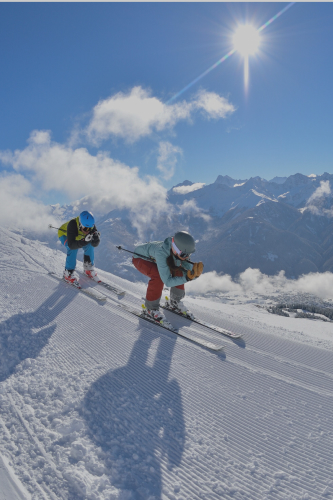 Zwei Skifahrer rasen auf einer sonnigen, verschneiten Piste mit blauem Himmel in Alpenresorts hinunter.