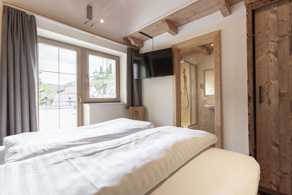 Gemütliches Schlafzimmer mit großem Fenster, TV, Holzakzenten und eigenem Bad mit Dusche.